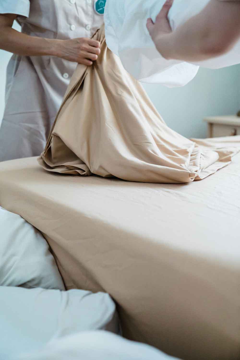 Camarera de pisos con uniforme colocando una sábana beige en la cama de una habitación de hotel
